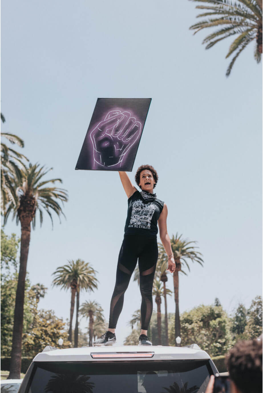 An activist standing on a car holding up a black sign with a purple outline of a fist