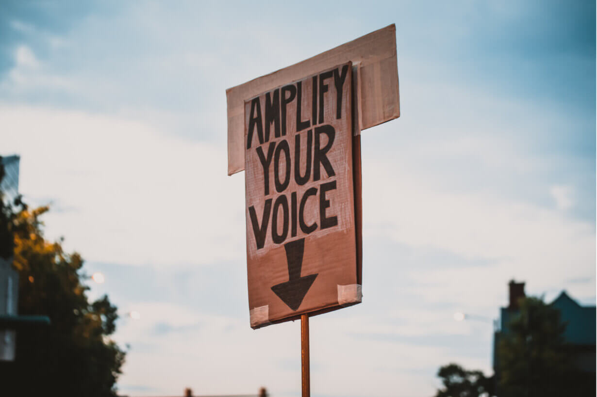a cardboard sign reading Amplify Your Voice with an arrow pointing down
