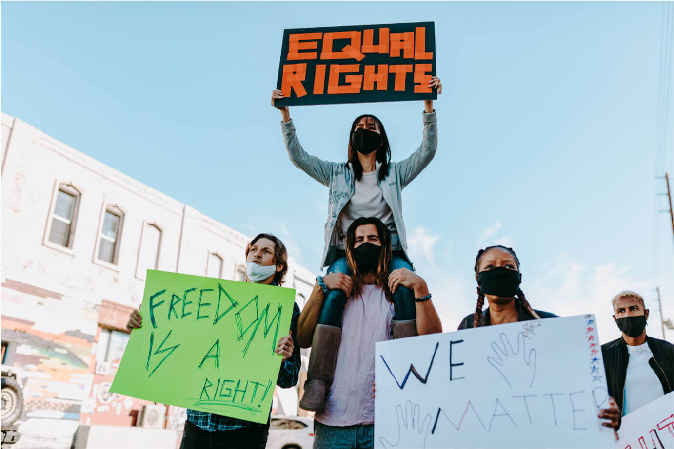 A photo taken at a protest; four people wearing medical masks, three of them holding signs. From left to right: a man holding a green handwritten Freedom is a human right sign; a woman sitting on a man's shoulders holding a black sign reading Equal Rights in orange block letters; a woman with braided hair holding a white We Matter sign