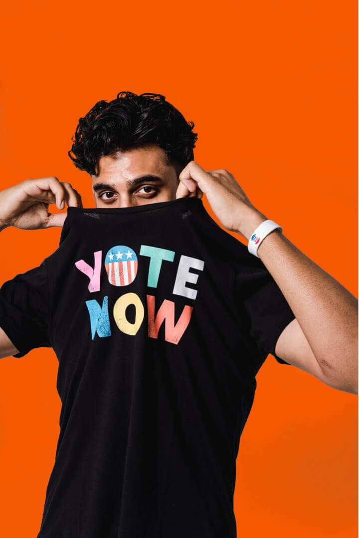 A man of color wearing a black Vote Now shirt and tugging it up over his mouth to emphasize the writing. He is standing in front of an orange backdrop.