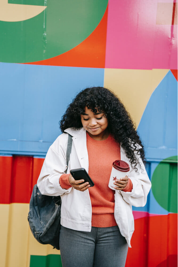 a black woman holding a coffee in one hand and looking at her phone in the other smiling. She's in front of a colorful background and has a bag slung over one shoulder.