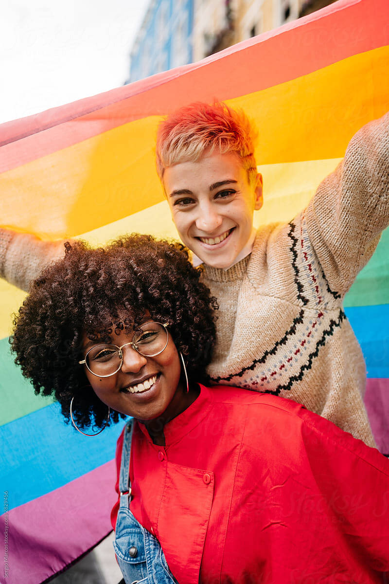 a black woman and white woman smiling and posing in front of a rainbow flag