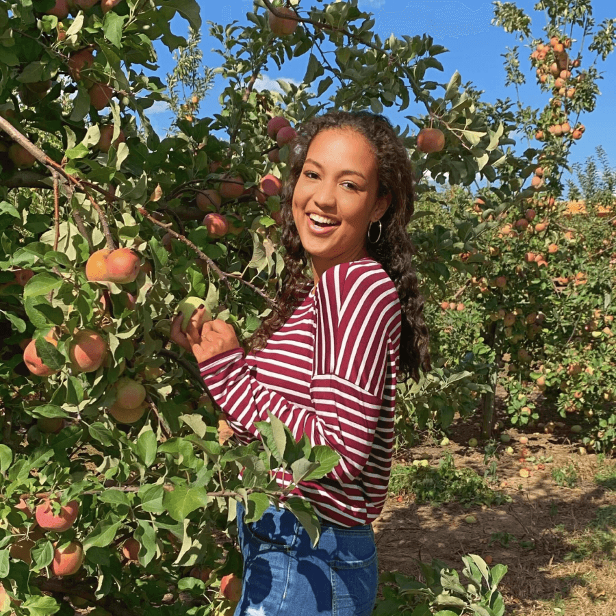 Maya wearing a white and red striped shirt and jeans and standing in an orchard grinning