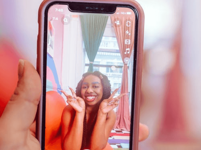 a phone with a photo of a black woman grinning holding up two peace signs in front of her windows. The phone is being held in a hand with the same backdrop as the photo, just much blurrier, behind it