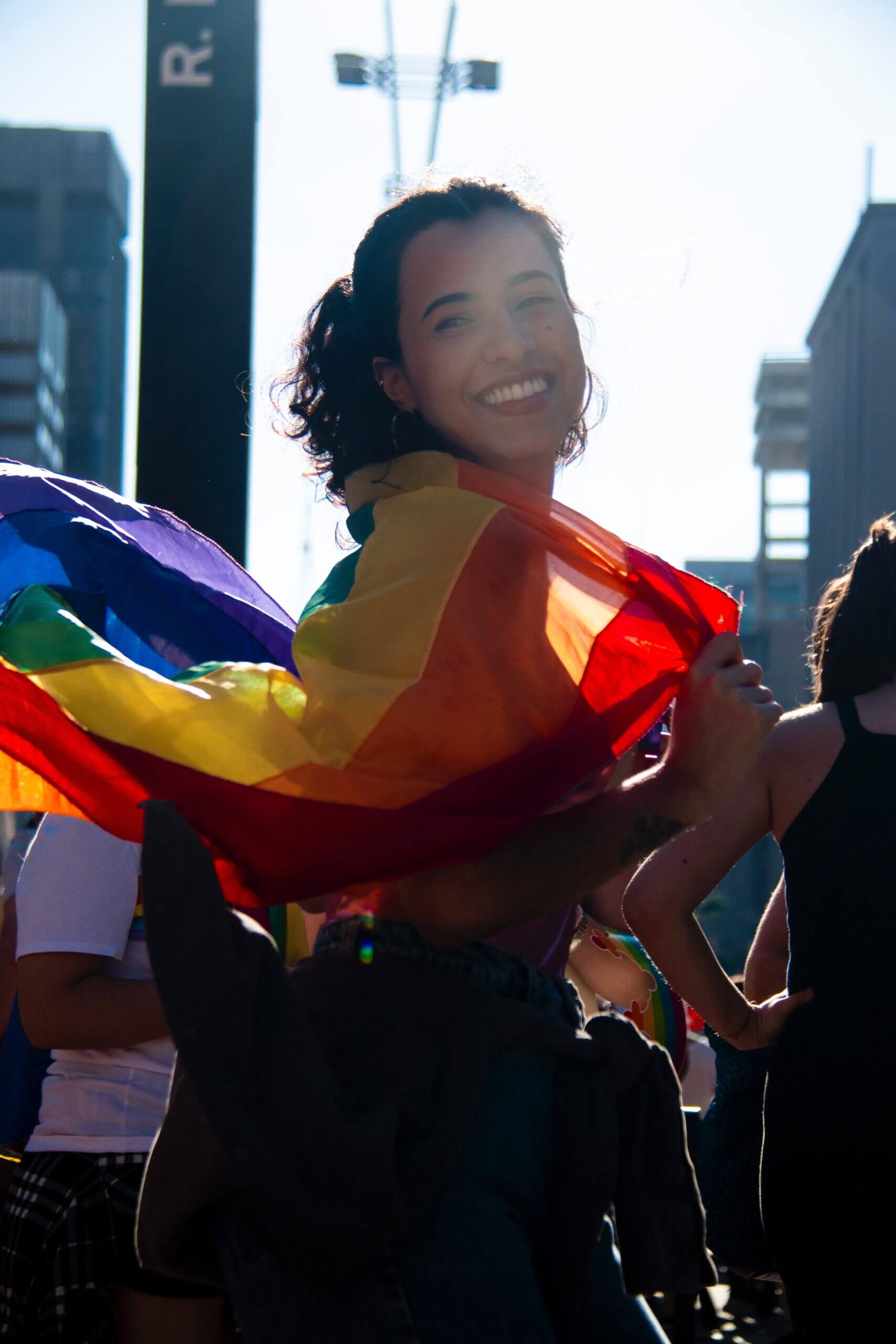 A woman of color wearing a rainbow flag around her shoulders and smiling at the camera while walking perpendicular from the camera
