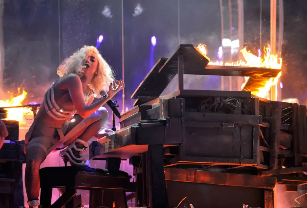 Lady Gaga in a nude body suit with piping at a piano mid-concert. The piano looks like it's on fire, one leg is up on the piano bench, her hand is curled in her signature "monster" hand gesture, and she's staring right at the camera.