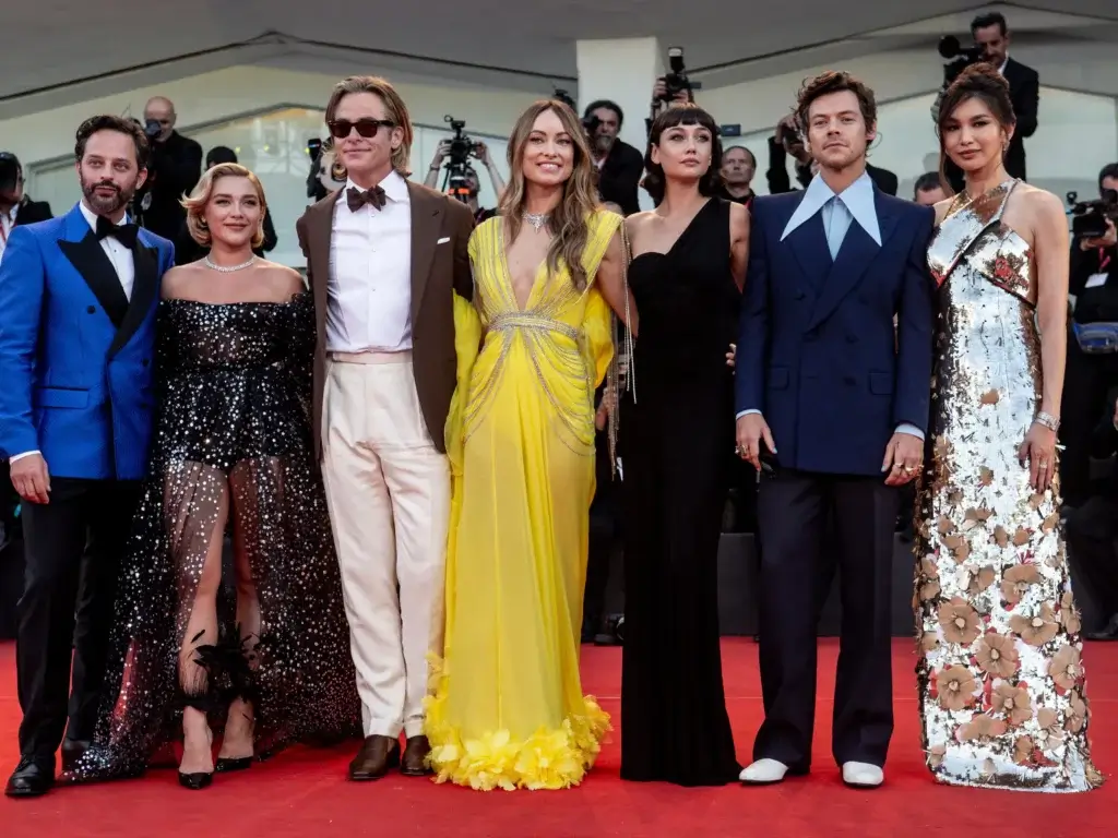 The Don't Worry Darling cast lined up at the movie premiere in Italy; in order from left to right it is Nick Kroll in a blue suit; Florence Pugh in a black romper; Chris Pine in cream and brown; Olivia Wilde in a yellow gown; Dakota Johnson in black; Harry Styles in blue; and Gemma Chan in a white floral gown