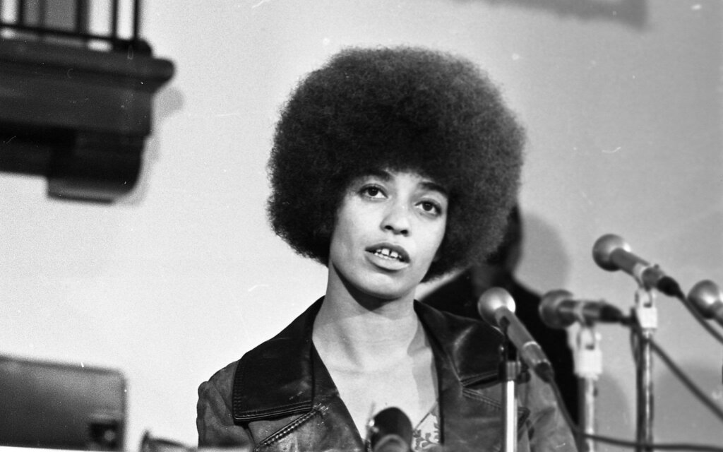 a black and white photo of Angela Davis from her early activism days. She is at a press conference with microphones in front of her, obviously mid-speech